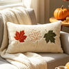 A cream-colored plush throw pillow featuring embroidered autumn leaves in shades of orange, brown, and green.