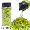 A glass jar of holographic chunky nail glitter mix with a black shaker top next to a pile of loose green and gold glitter.