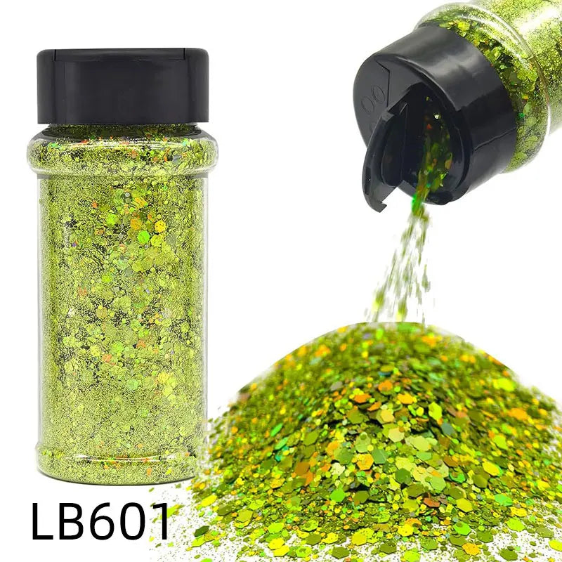 A glass jar of holographic chunky nail glitter mix with a black shaker top next to a pile of loose green and gold glitter.