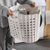 A person is holding a white folding laundry basket with handles, featuring perforated sides, in a domestic setting.
