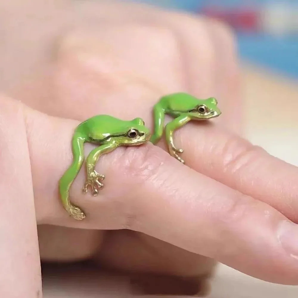 A hand wearing a ring shaped like a small green tree frog, featuring bright green enamel and gold accents.