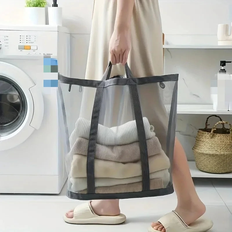 A person holds a black mesh laundry basket with a handle, filled with folded white and beige clothing.