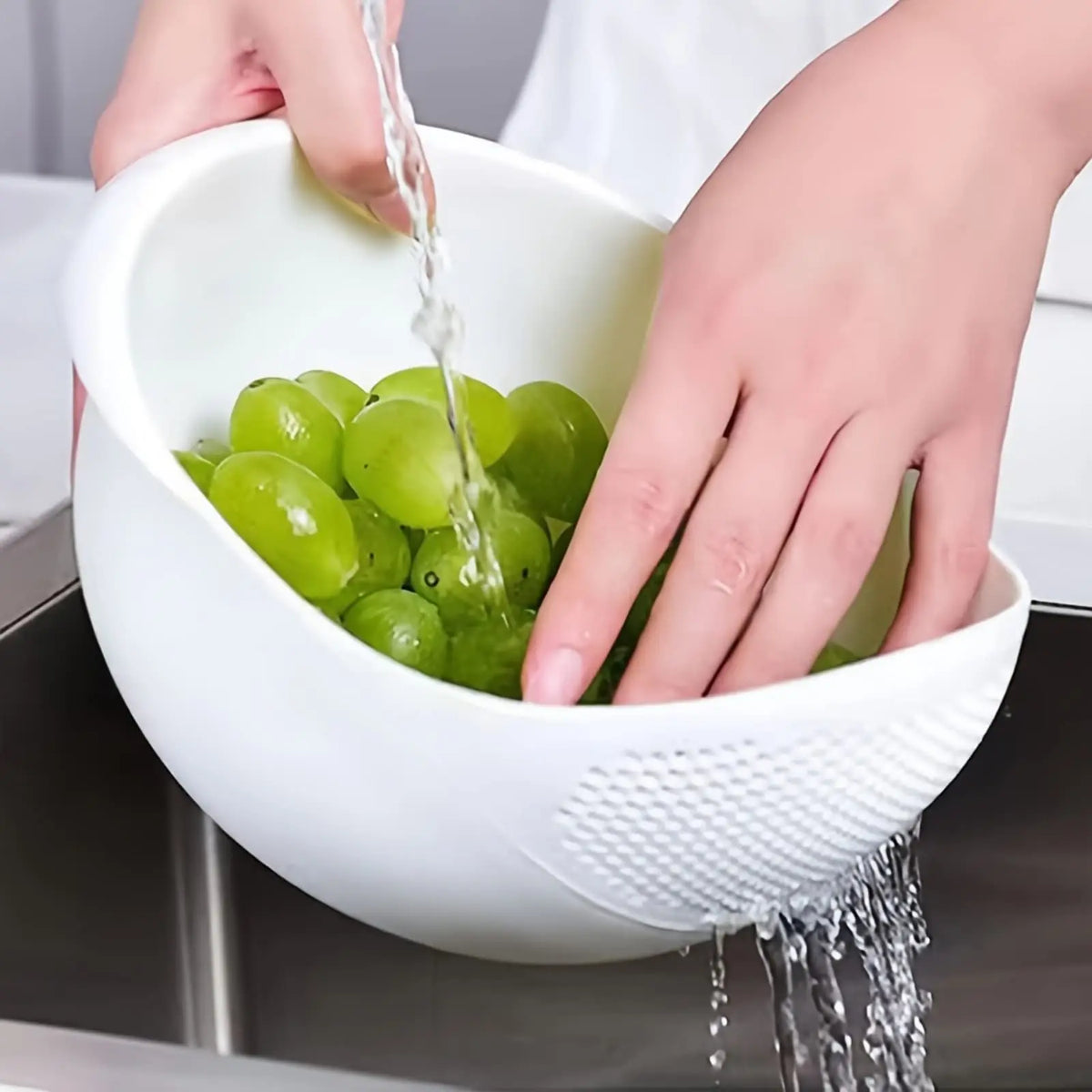 Hands are pouring water into a white multi-purpose kitchen wash basket filled with green grapes.