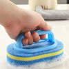 A hand holds a blue sponge cleaning brush with a yellow band on a white surface.
