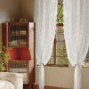 A set of sheer white floral lace curtains hanging in a sunlit room with wooden cabinets and large windows.