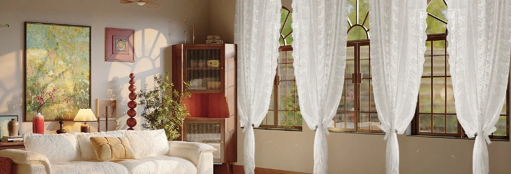A set of sheer white floral lace curtains hanging in a sunlit room with wooden cabinets and large windows.