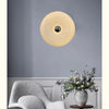 A modern, round, cream-colored LED wall sconce is mounted on a gray wall above a white upholstered chair.