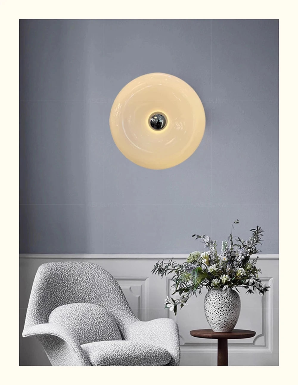 A modern, round, cream-colored LED wall sconce is mounted on a gray wall above a white upholstered chair.