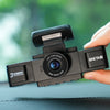 A hand holds a black 3-channel dash cam with a wide-angle lens, set against a blurred green outdoor background.