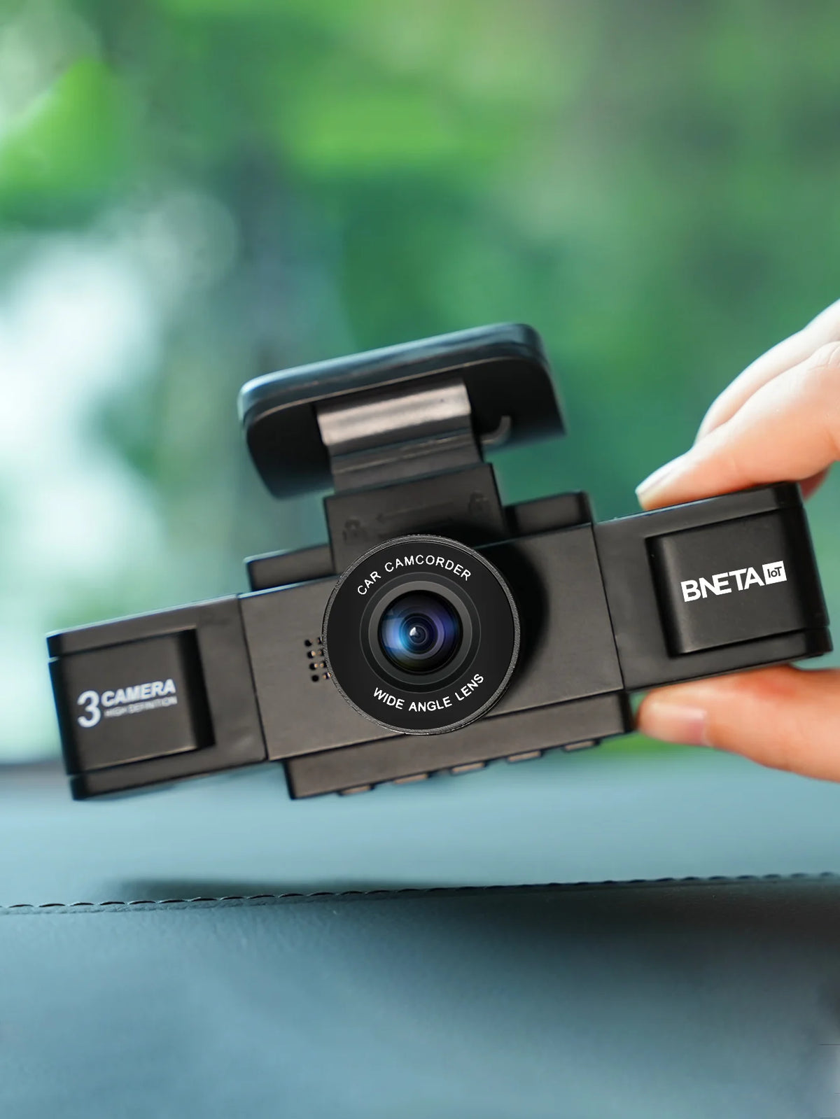 A hand holds a black 3-channel dash cam with a wide-angle lens, set against a blurred green outdoor background.
