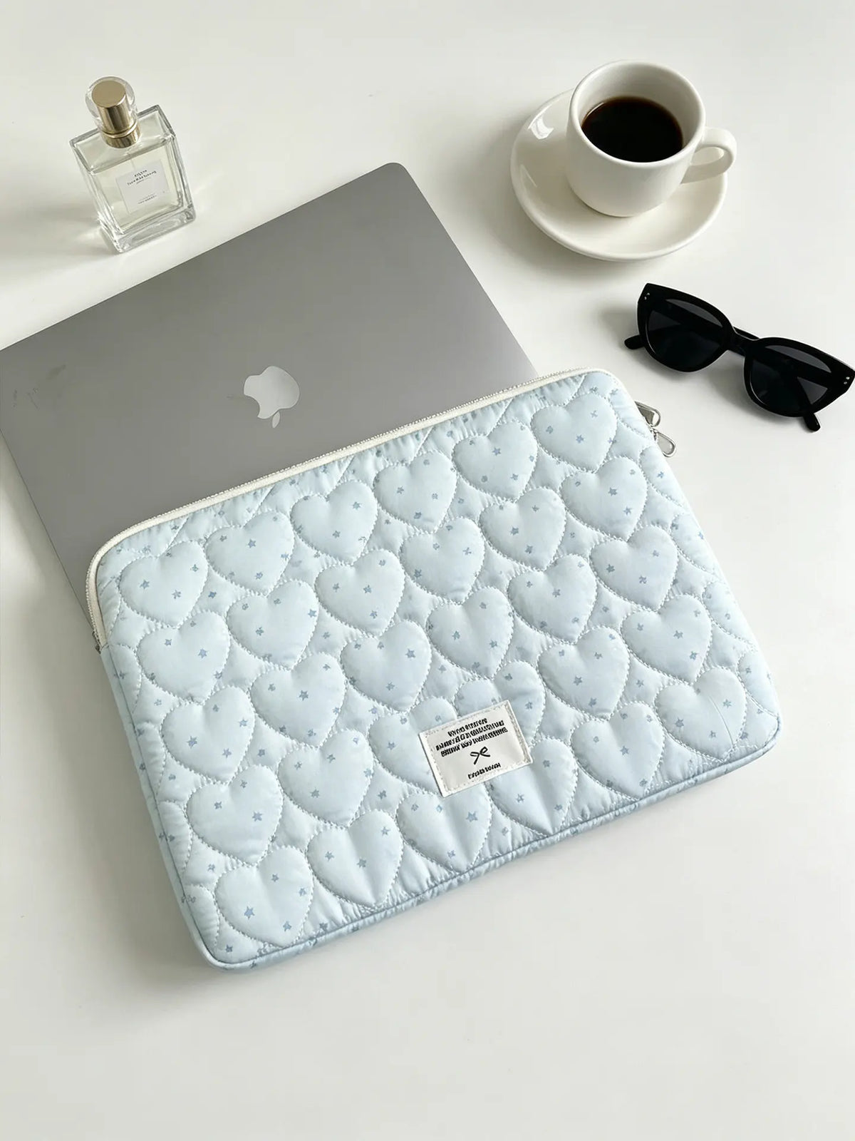 A light blue quilted laptop sleeve is placed next to a silver laptop and sunglasses on a white surface.