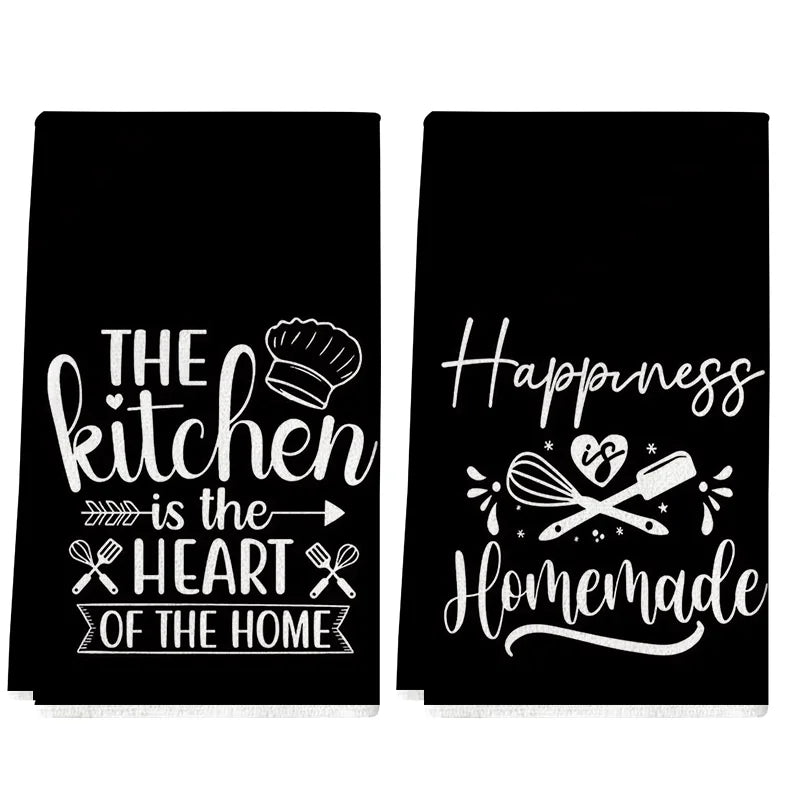 A set of two black kitchen towels featuring white text slogans like The kitchen is the heart of the home and Happiness Homema