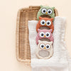 A set of silicone owl-shaped teethers and rattles in various pastel colors resting on a woven tray and white cloth.