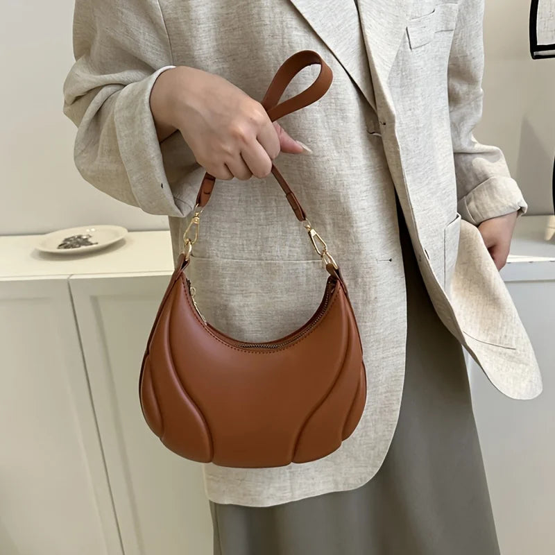 A woman holds a brown leather crossbody bag with a gold chain strap against a light beige blazer.