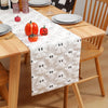 White table runner with black ghost pattern and autumn pumpkins on a wooden table.