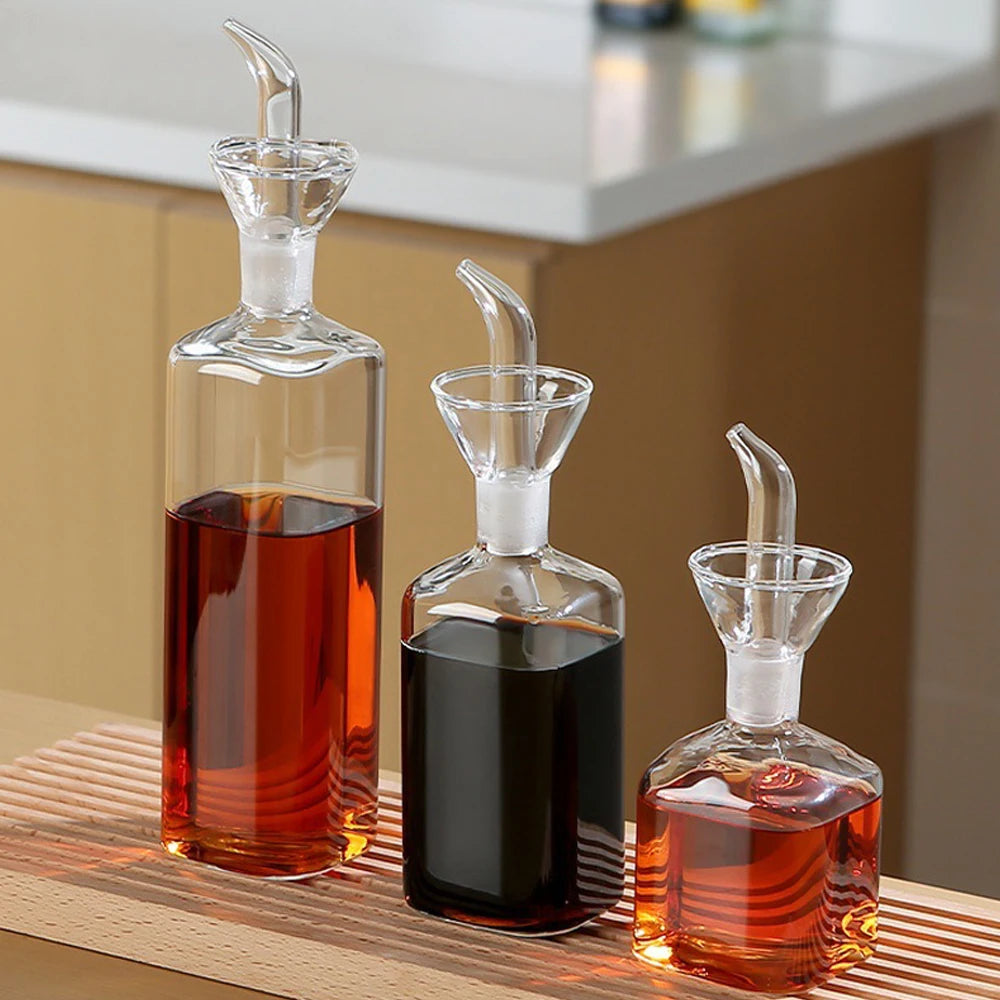 A clear borosilicate glass oil and vinegar dispenser with a long spout is shown on a wooden surface.