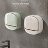 A close-up of a modern, flip-top soap holder in light green and beige tones mounted on a gray wall above a sink.
