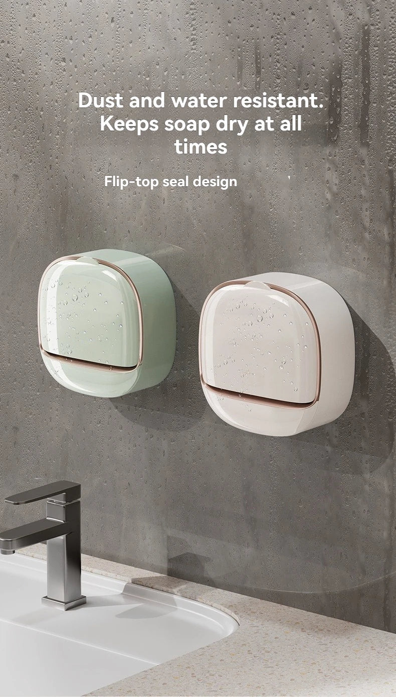 A close-up of a modern, flip-top soap holder in light green and beige tones mounted on a gray wall above a sink.