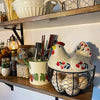 A rustic ceramic hen-shaped egg basket with a lid sits on a wooden shelf among various kitchen items.
