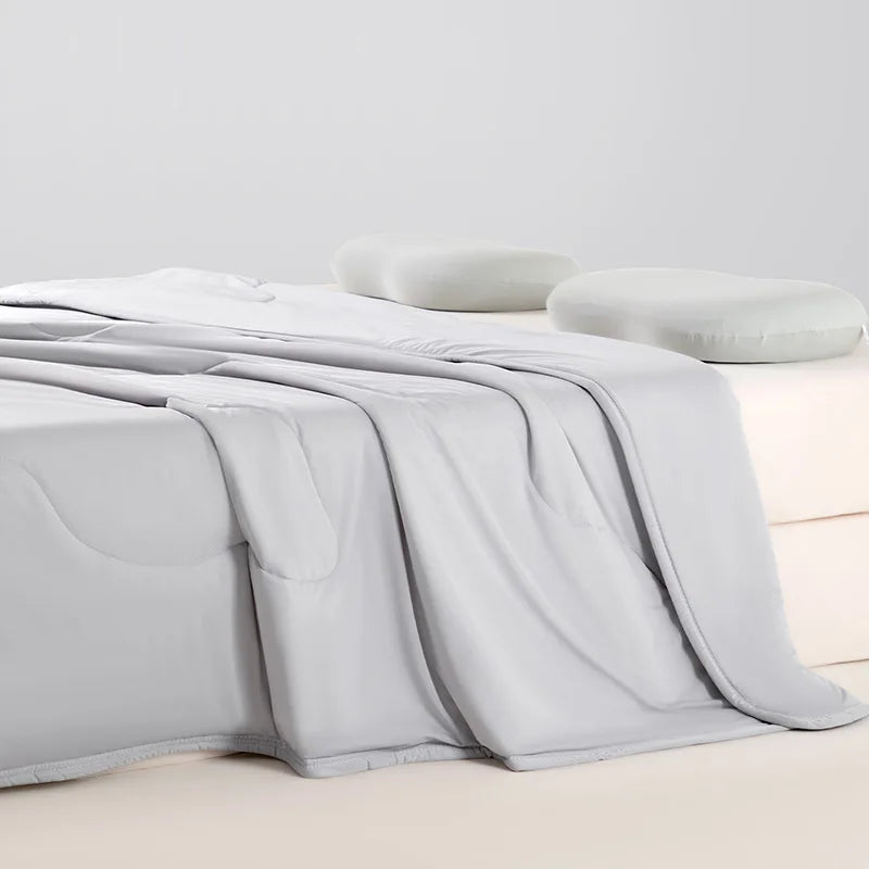 A light gray ice silk cooling blanket is draped over a white surface next to a soft pillow.
