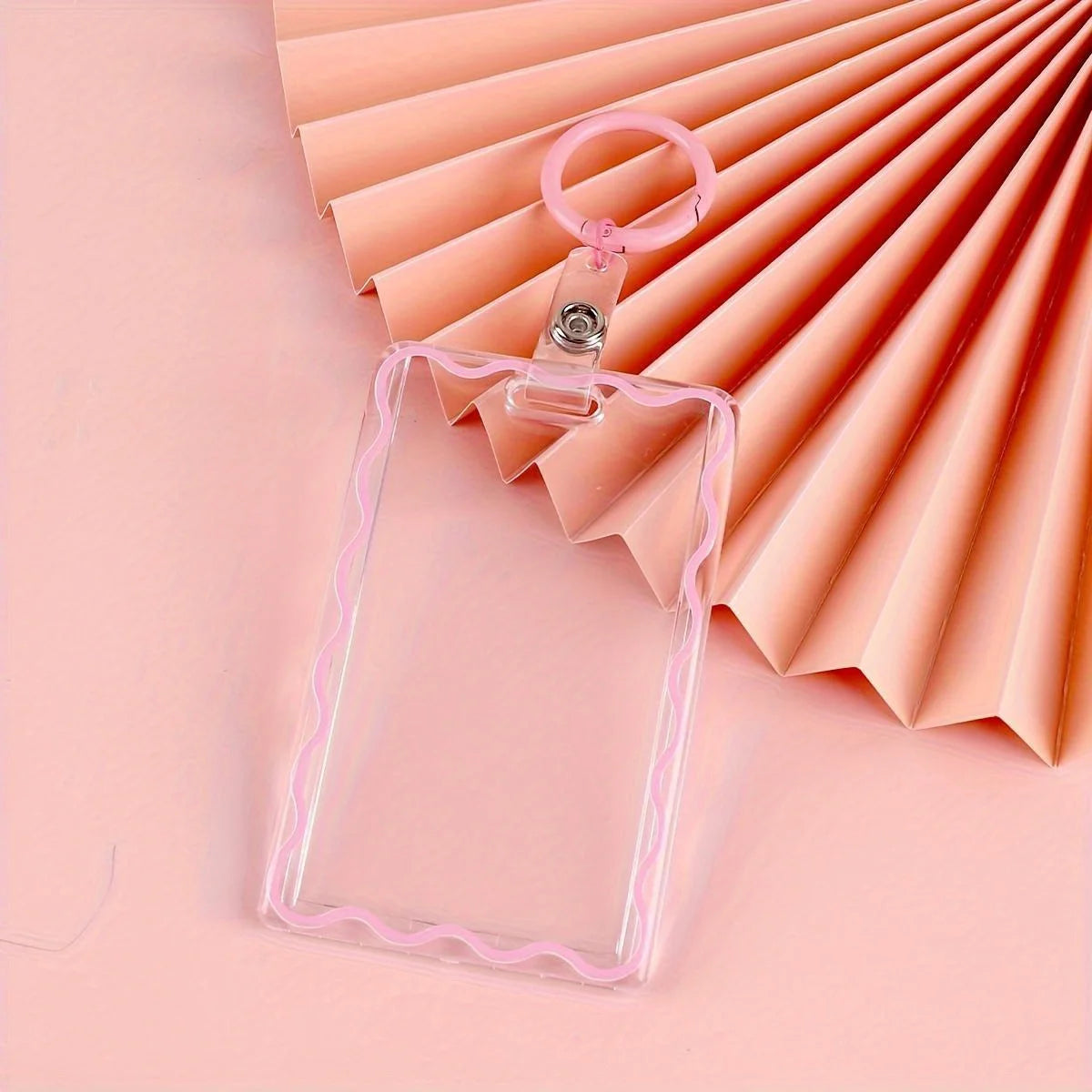 Sujetador de tarjetas de acrílico transparente con borde rosa y llavero, sobre fondo de papel plisado rosa.