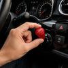A hand is pressing a red engine start button cover on a car dashboard control panel.