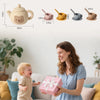 A woman smiles while holding a pink silicone bear teapot set for toddlers in a bright living room setting.