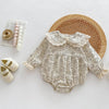 A cream floral lace cotton romper for a baby girl displayed on a white surface with a woven mat.