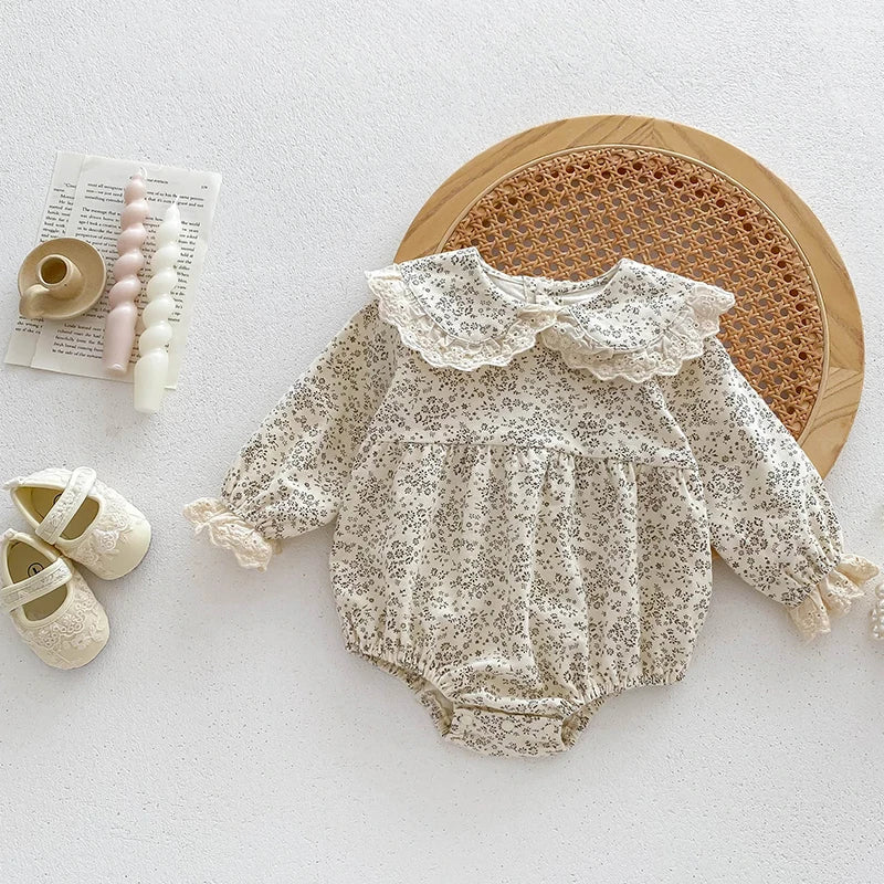 A cream floral lace cotton romper for a baby girl displayed on a white surface with a woven mat.