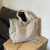 A beige corduroy tote bag with a light strap resting on a wooden surface next to a dark cushion.