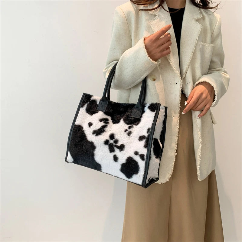 A woman holds a black and white faux fur leopard print tote bag in front of a light beige blazer.