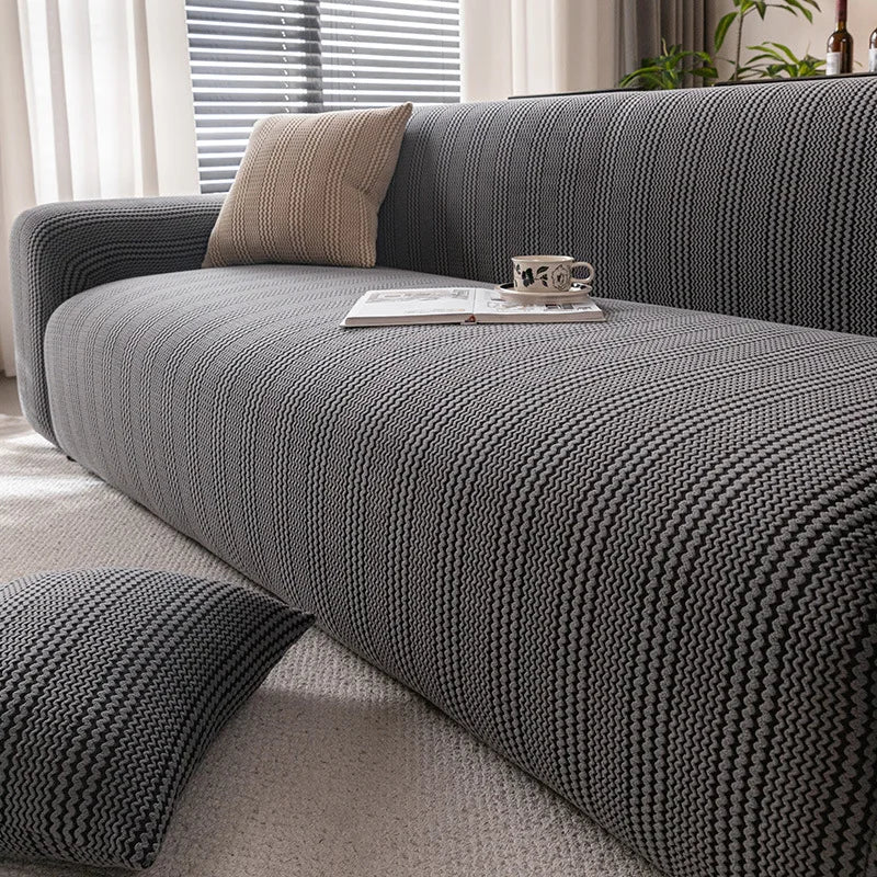 A dark gray quilted stretch sofa cover with a textured weave and a beige throw pillow on a light rug.