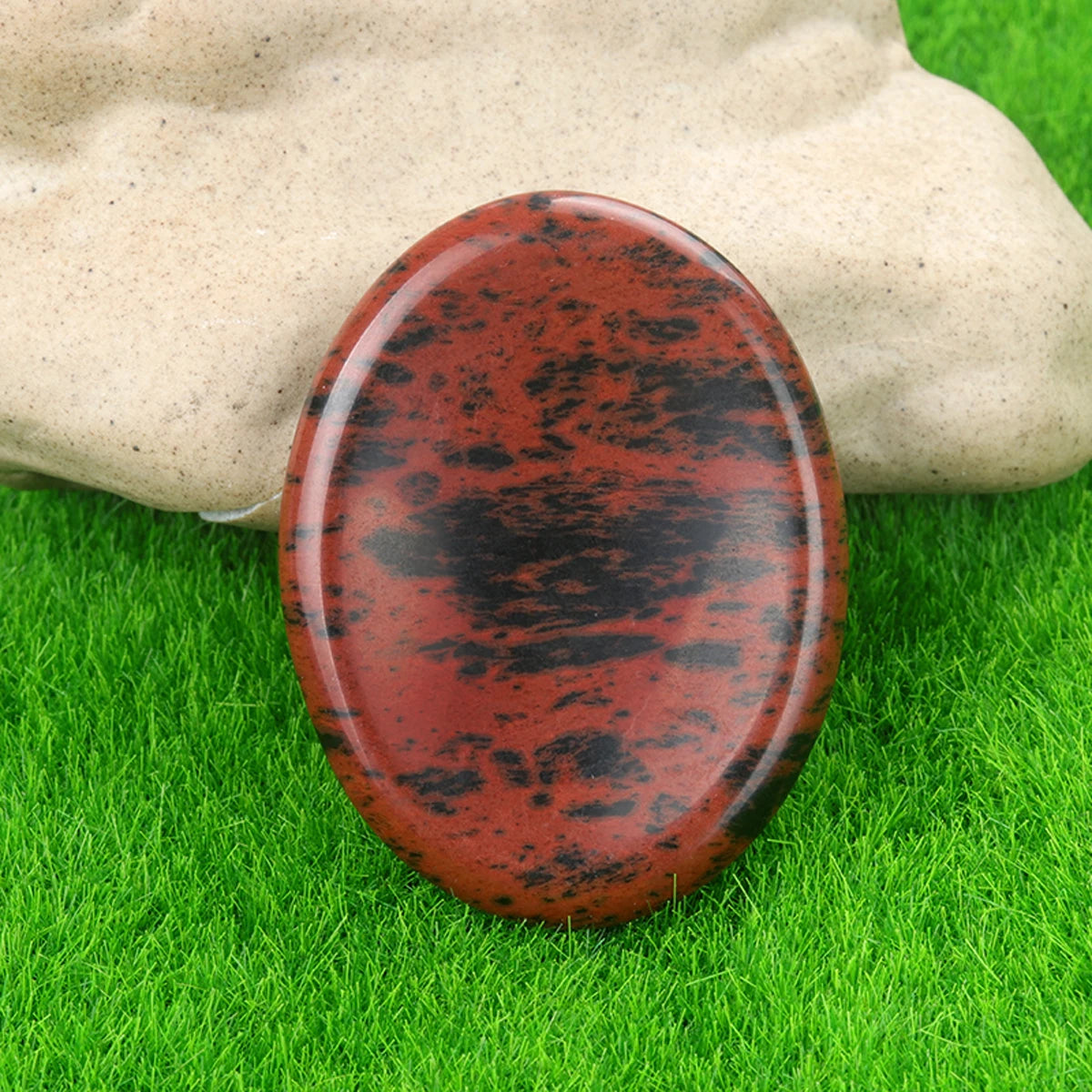 A reddish-brown oval natural crystal stone massage stone resting on green grass.