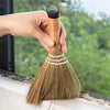 A hand holds a natural grass broom with a wooden handle and straw bristles, positioned near a window sill.