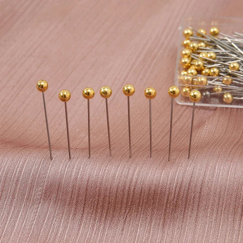 A collection of gold-colored precision quilting pins with silver shafts is displayed on a pink fabric.