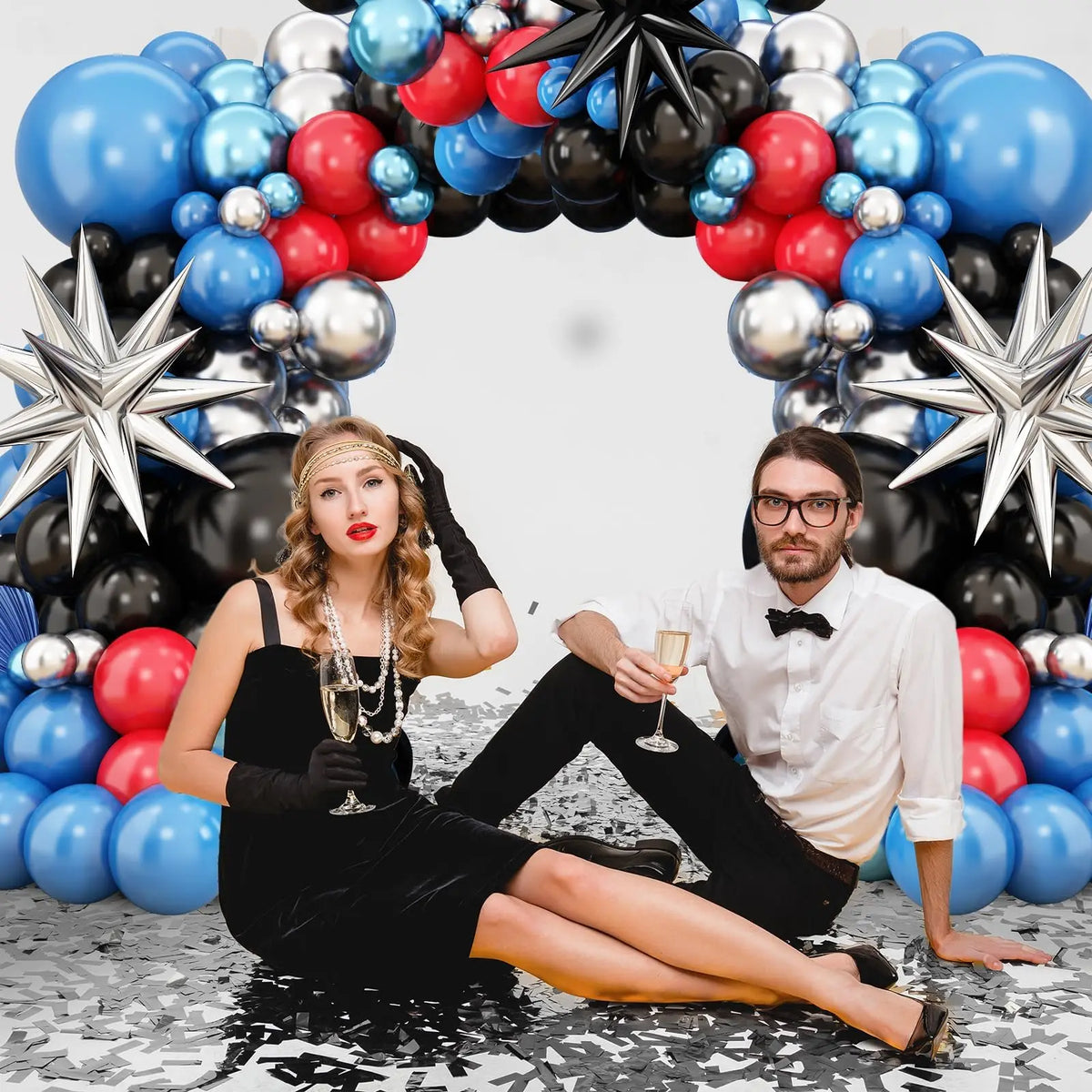 A woman in a black dress lies on the ground next to a man, surrounded by a balloon garland arch of blue, red, and black ballo