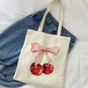 A canvas tote bag featuring a pink cherry print on a light beige background, resting on a white sheet.