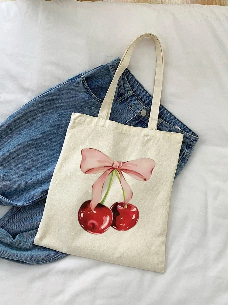 A canvas tote bag featuring a pink cherry print on a light beige background, resting on a white sheet.