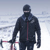 A person wearing a black windproof full face balaclava mask and dark clothing is riding a bicycle in a snowy outdoor setting.