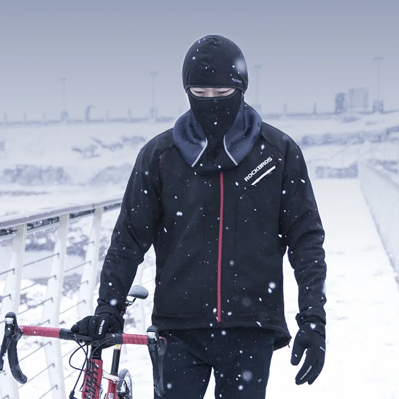 A person wearing a black windproof full face balaclava mask and dark clothing is riding a bicycle in a snowy outdoor setting.