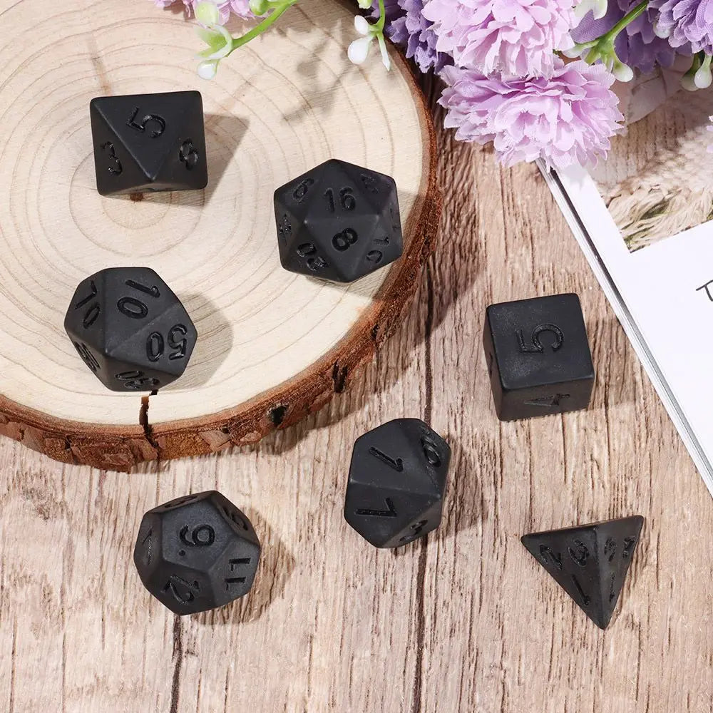 A set of matte black polyhedral dice resting on a wooden surface next to purple flowers.
