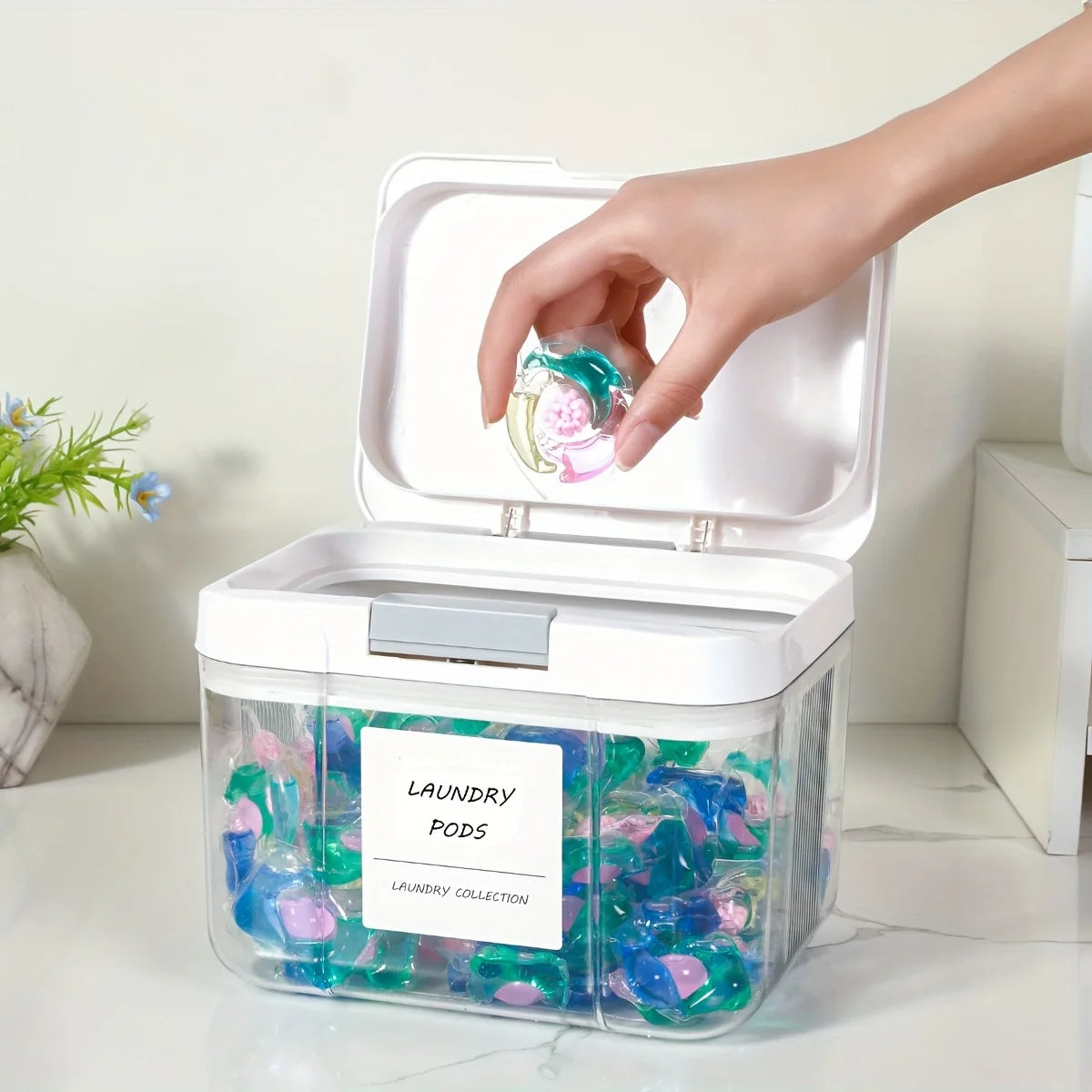 A hand is opening an automatic pop-up laundry pod holder with an airtight lid, revealing colorful laundry pods inside.