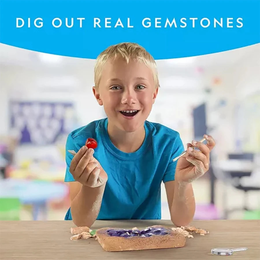 A young boy holds a small piece of rock while looking at a wooden tray with crystals and tools for a gemstone digging kit.
