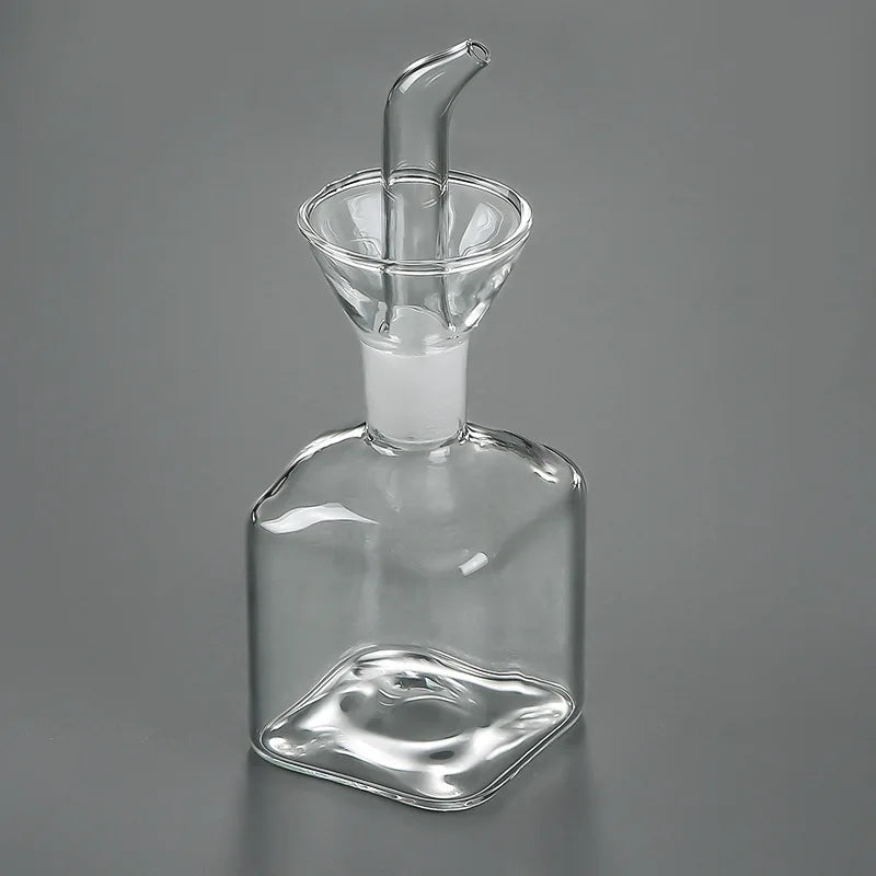 A clear borosilicate glass oil and vinegar dispenser with a spout and a curved base.