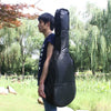 A man stands outdoors holding a black acoustic guitar soft case with a padded shoulder strap.