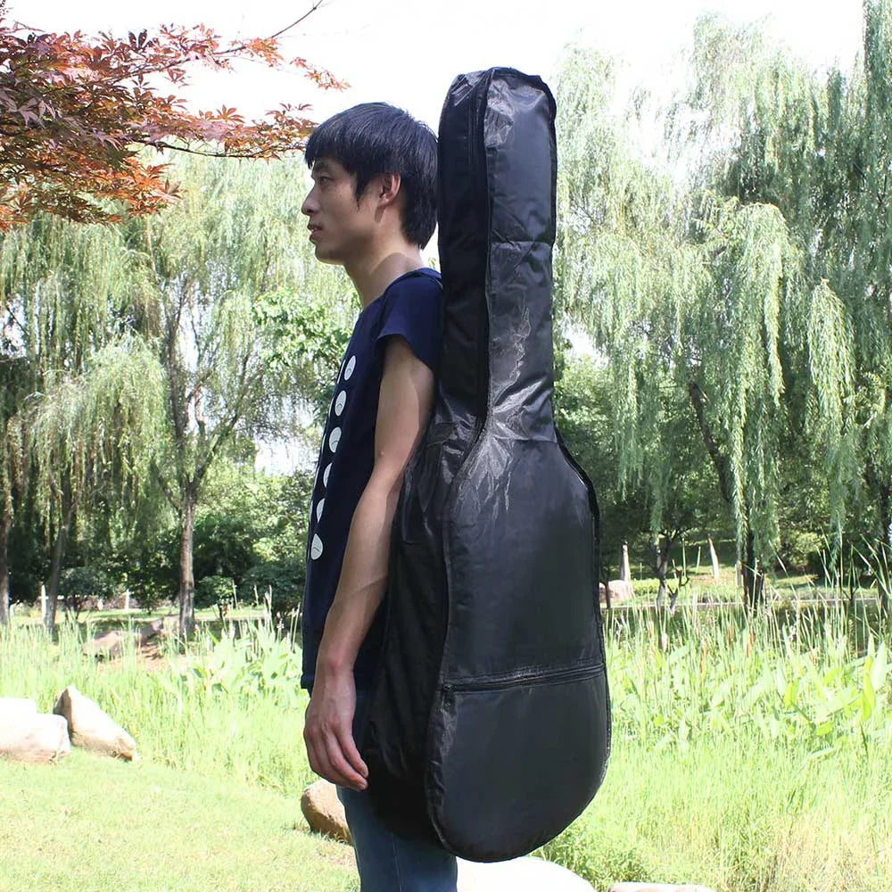 A man stands outdoors holding a black acoustic guitar soft case with a padded shoulder strap.