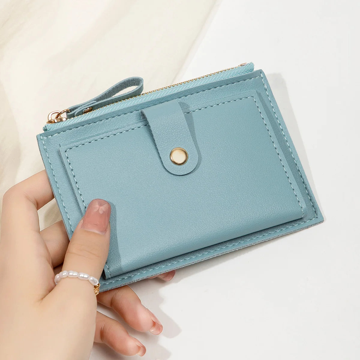 A hand holds a light blue slim PU leather card holder wallet with a gold zipper and a round button.