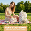A smiling woman sits on the grass with a floral backpack, handbag, and pencil case, ready for a picnic.