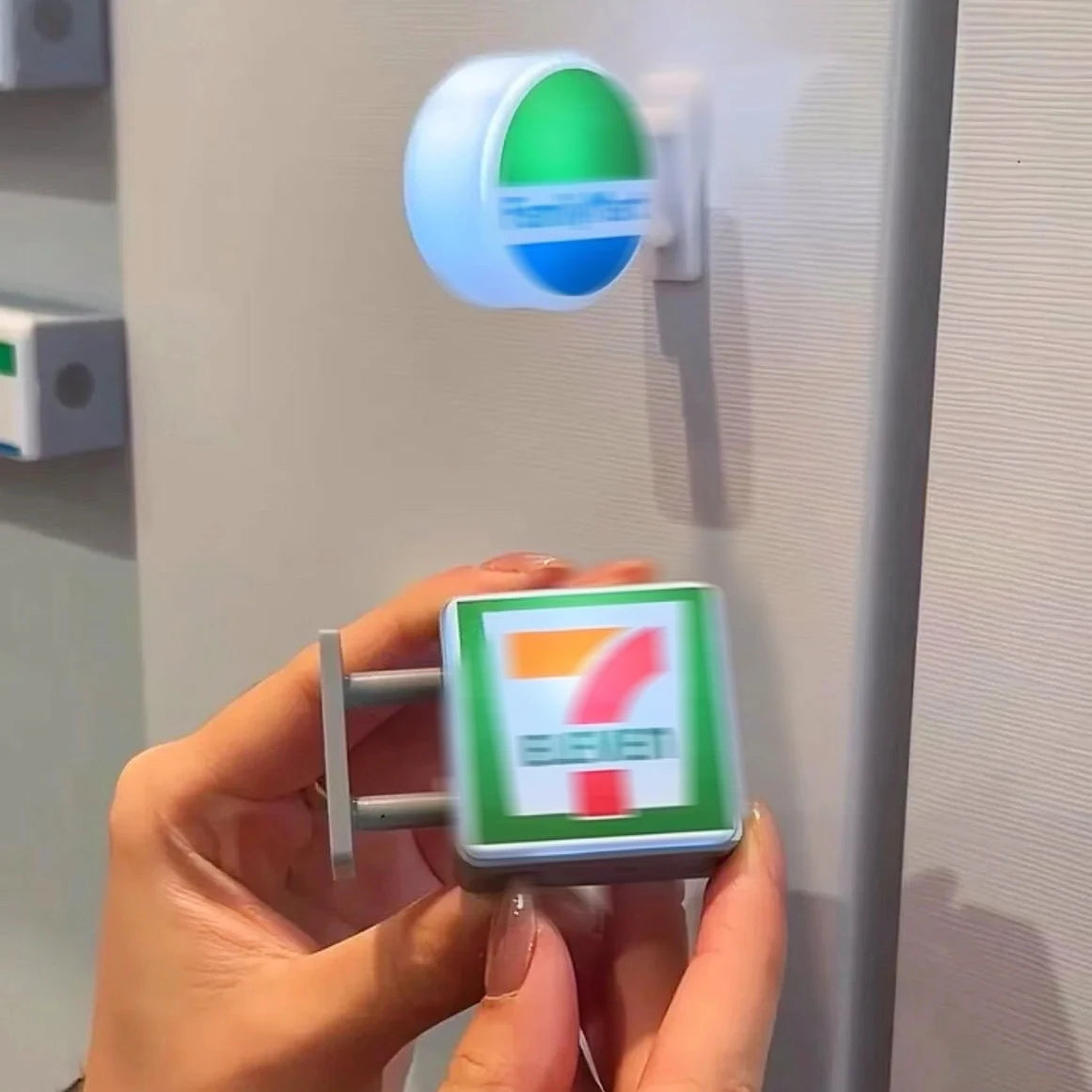 A hand holds a miniature glowing storefront fridge magnet with a red number and green background against a white refrigerator
