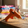 An orange hot melt glue gun with a stand and glue sticks resting on a wooden surface next to various craft supplies.